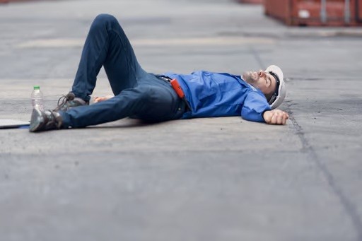 A person wearing a hard hat and blue shirt is lying on their back on a concrete surface, appearing unwell or injured.