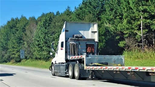 A white semi-truck with a flatbed trailer drives on a highway next to a speed limit sign and a backdrop of green trees.