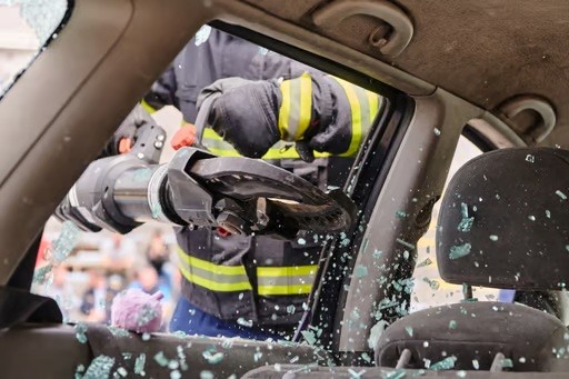 A firefighter uses hydraulic rescue tools to cut through a car door window, with glass shards flying inside the vehicle.