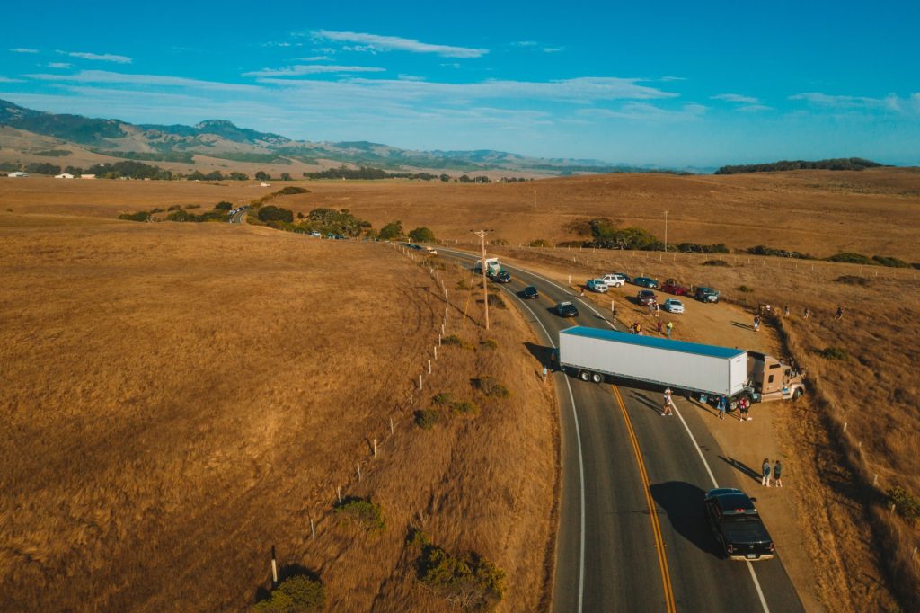 Aerial view of a semi-truck stopped on the side of a rural road with several cars and people nearby in a dry, open landscape.