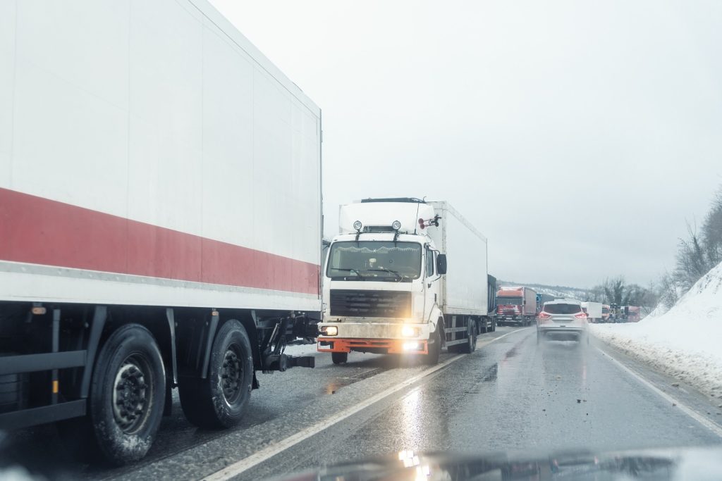 Two large trucks pass each other on a wet, snow-lined road, with cars and more trucks visible ahead under overcast skies.