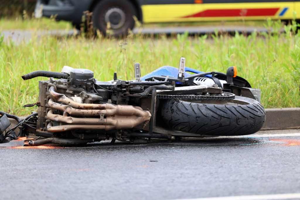 A motorcycle lies on its side on a wet road near a grassy area, with an emergency vehicle blurred in the background.