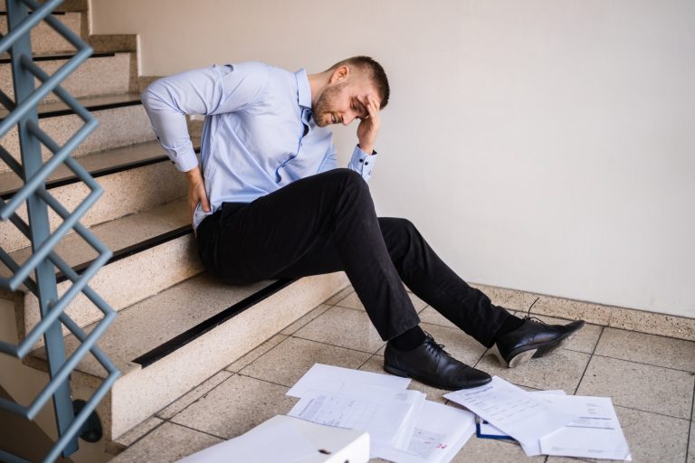 A man in business attire sits on stairs holding his lower back, appearing in pain, with scattered papers and folders on the floor around him.
