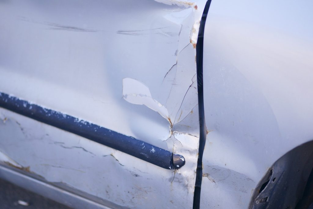 Close-up of a white car door with significant dents, scratches, and paint damage, indicating it has been involved in a collision.