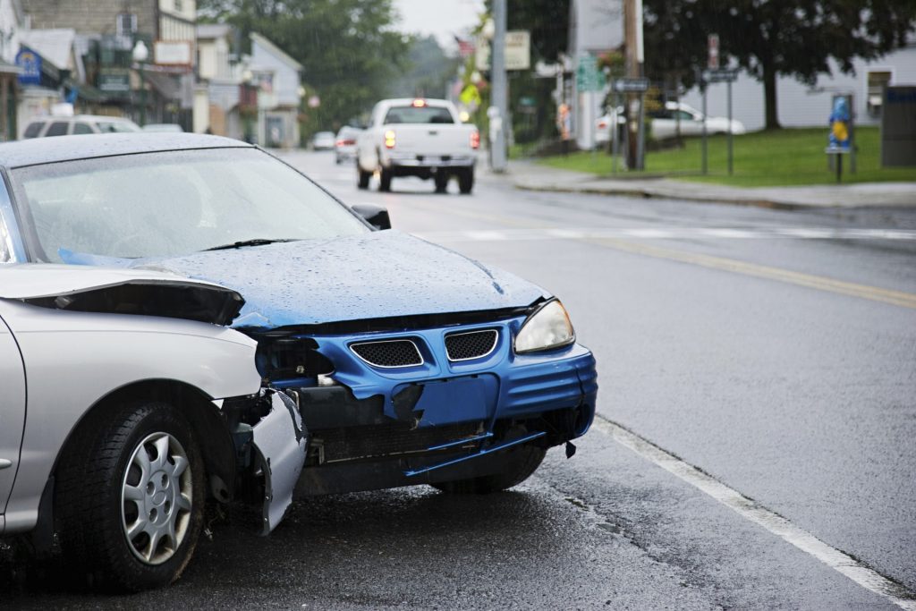 A silver car with a damaged front bumper and hood is stopped at an intersection on a wet street in a small town.