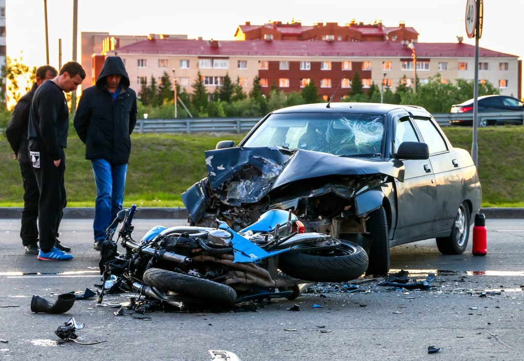 A heavily damaged car and motorcycle after a collision, with three men standing nearby on the road in front of apartment buildings.