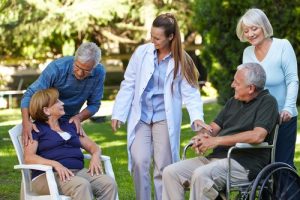 A doctor talks with four senior adults, two seated and two standing, outdoors in a garden setting. One man uses a wheelchair, and the group appears to be engaged in friendly conversation.