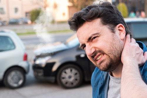 Man standing outdoors holding his neck in apparent pain with two cars in the background that appear to have been in a collision.