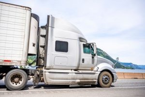 A silver semi-truck with a trailer is parked on the side of a highway, with a river and green hills visible in the background.