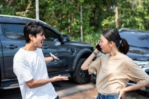 A man gestures and talks to a woman on the phone in front of two damaged cars after an apparent accident on a street with trees in the background.