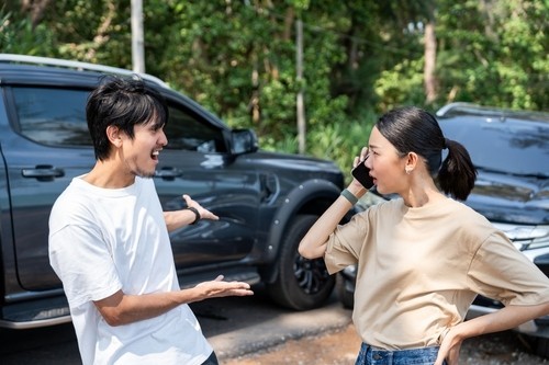 A man gestures and talks to a woman on the phone in front of two damaged cars after an apparent accident on a street with trees in the background.