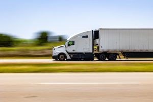 A white semi-truck with a trailer drives on a highway during the day, with a blurred background indicating motion.