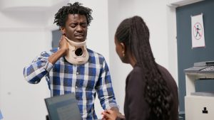 A man wearing a neck brace holds his neck in discomfort while speaking to a woman at an office desk.