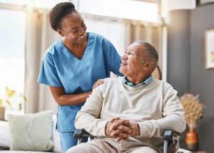 A nurse in blue scrubs stands beside an older man sitting in a wheelchair, both smiling at each other in a well-lit room.
