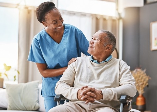 A nurse in blue scrubs stands beside an older man sitting in a wheelchair, both smiling at each other in a well-lit room.