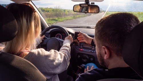 Two people sit in the front seats of a car; the driver holds the steering wheel, while the passenger points at the dashboard.