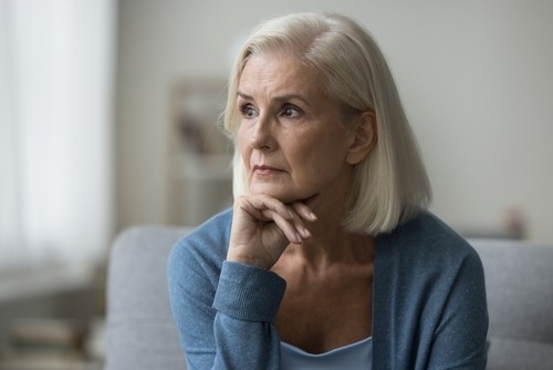 Older woman with short gray hair sits indoors, resting her chin on her hand and looking thoughtfully into the distance.