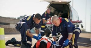 Two paramedics provide emergency medical assistance to a person lying on the ground near an ambulance with its rear doors open.