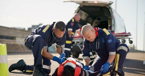 Two paramedics provide emergency medical assistance to a person lying on the ground near an ambulance with its rear doors open.