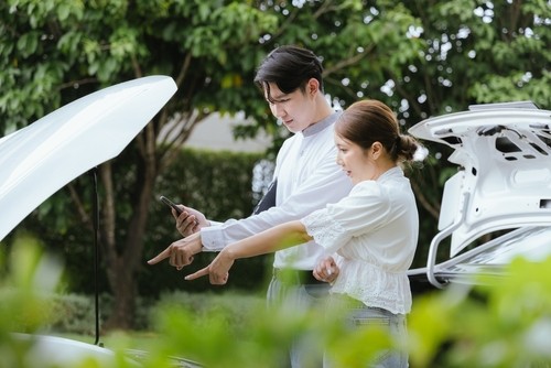 A man and woman stand beside a car with the hood open, both pointing and looking at the engine while the man holds a phone.