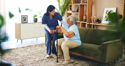 A healthcare worker assists an older woman with a cane as she sits on a green couch in a living room.