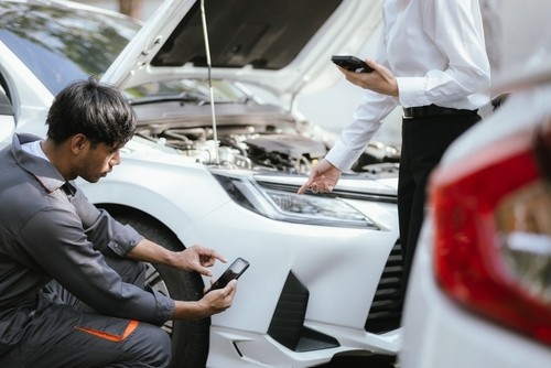 Two people inspect the front of a white car with the hood open; one crouches and takes a photo with a smartphone while the other stands and points.