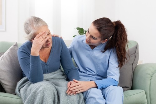 A nurse in blue scrubs sits beside an older woman with a blanket, comforting her as she holds her head in distress.