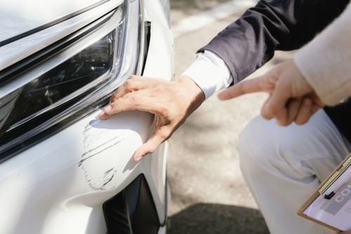 Two people inspect a white car with visible scratches on the front bumper; one person points at the damaged area.
