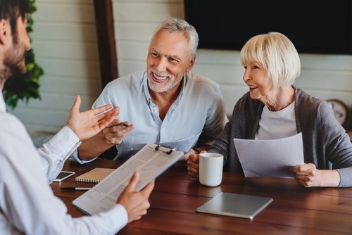 Smiling older couple sits at a table holding papers and a mug, listening to a person with a clipboard during a meeting or consultation.