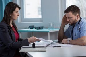 A woman in business attire shows documents to a male healthcare worker in scrubs, who appears stressed, in an office setting.