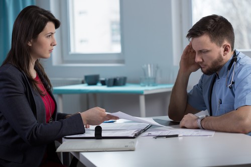 A woman in business attire shows documents to a male healthcare worker in scrubs, who appears stressed, in an office setting.