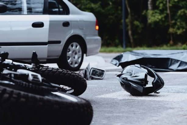 A damaged motorcycle, helmet, and side mirror lie on the road near a silver car, with a black body bag in the background.