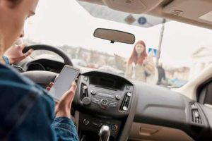 A driver is looking at a smartphone while approaching a pedestrian who is crossing the street in front of the car.