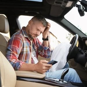 A man sitting in the driver’s seat of a car looks at his phone with a confused expression, one hand scratching his head.