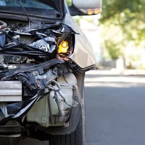 Front end of a white car severely damaged in a collision, with exposed internal parts and a headlight still on, parked on a street.