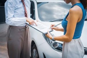 Two people stand near a car, one pointing at a document while the other holds a pen, possibly discussing or signing an agreement.