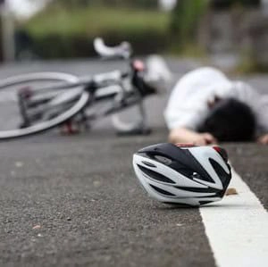 A bicycle and a person lie on the ground with a helmet in the foreground, suggesting a recent accident on a paved road.