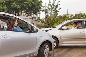 Two cars in a minor accident on a road. A woman in one car and a man in the other car look at each other. Trees and a fence are in the background.