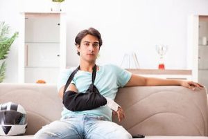 A young man with his right arm in a black sling sits on a beige couch, with a motorcycle helmet beside him and a trophy in the background.