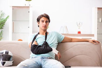 A young man with his right arm in a black sling sits on a beige couch, with a motorcycle helmet beside him and a trophy in the background.