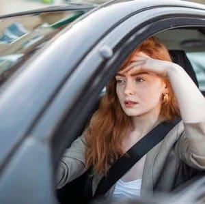A woman with long red hair sits in a car driver's seat, wearing a seatbelt and looking out the window with her hand on her forehead.