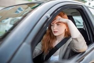 A woman with long hair sits in a car, wearing a seatbelt, resting her head on her hand while looking out the window.