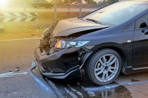 Front view of a black car with significant damage to the hood and bumper, parked on an asphalt road near a guardrail.