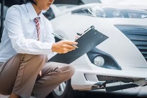 Person inspecting the damage on a white car's bumper with a clipboard and pen.