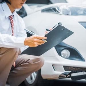A person in business attire kneels near a white car, holding a clipboard and pen, appearing to inspect or assess the vehicle.