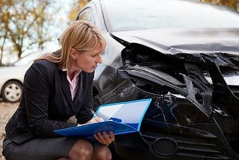 A woman in business attire examines and takes notes on a damaged car with a crumpled front end.