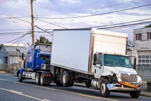 A white damaged box truck is being towed by a blue semi-truck on a road with industrial buildings in the background.