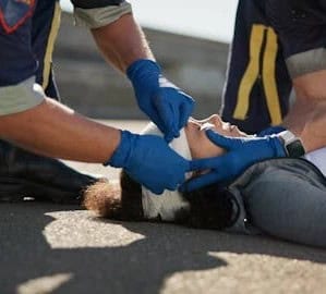 Two emergency responders wearing blue gloves are wrapping a bandage around the head of an injured person lying on the ground.