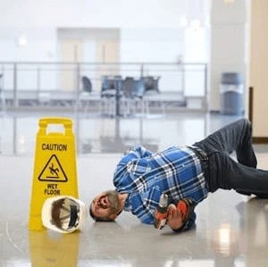 A man in a plaid shirt lies on the floor next to a "Caution: Wet Floor" sign, appearing to have slipped and fallen.