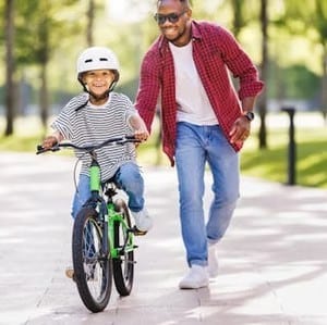 An adult helps a smiling child wearing a helmet ride a green bicycle on a sunny path lined with trees.